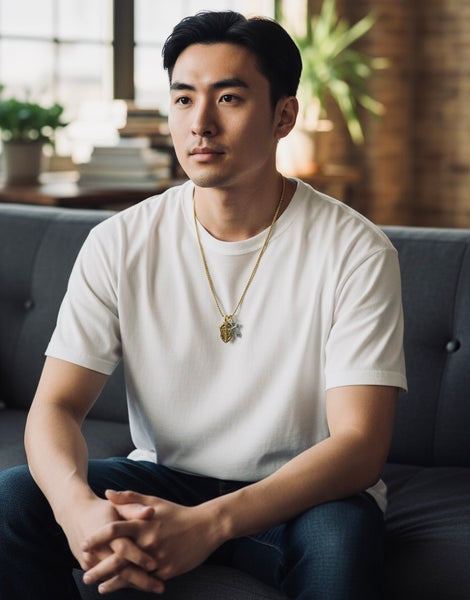 Man sitting on a couch wearing a white t-shirt and gold and silver shield and cross necklace in a cozy room.