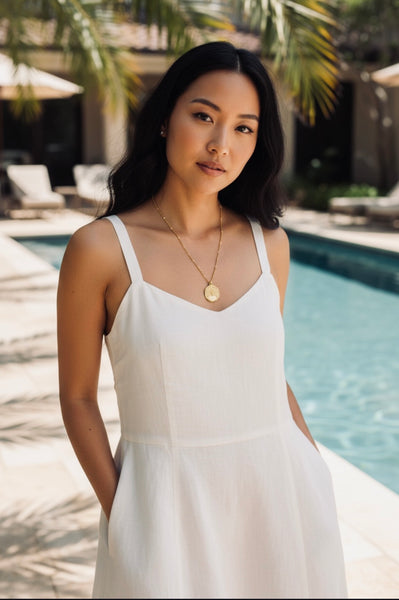Woman in a white dress wearing a gold peacock pendant necklace standing by a pool with palm trees in the background