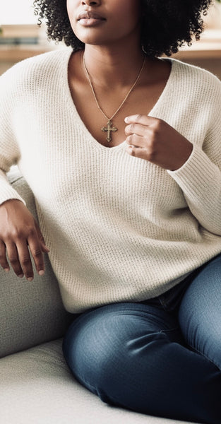 Person wearing a white sweater and blue jeans with a large gold cross necklace, sitting on a couch.