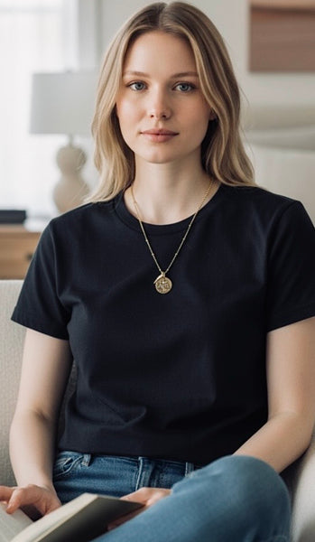 Woman wearing a religious alpha omega coin necklace on a satellite chain and a black t-shirt and blue jeans sitting on a couch with a book.