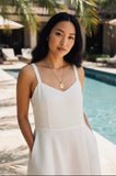 Woman in a white dress wearing a gold peacock pendant necklace standing by a pool with palm trees in the background