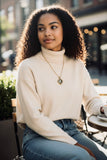 Woman sitting outdoors wearing a cream sweater and blue jeans, with a gold and silver lion coin in holder pendant necklace
