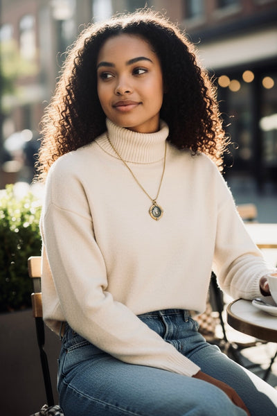 Woman sitting outdoors wearing a cream sweater and blue jeans, with a gold and silver lion coin in holder pendant necklace