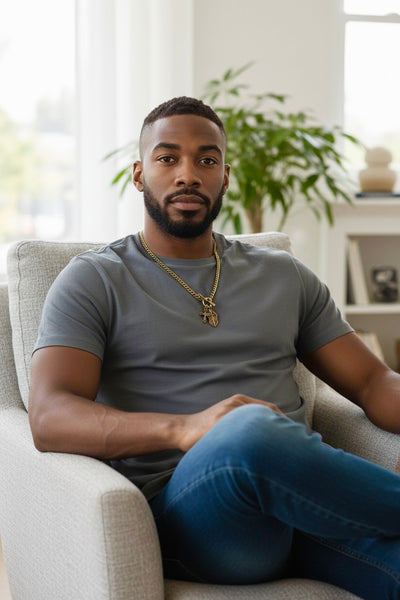Man sitting on a couch in a living room wearing a heavy gold necklace with a faith inspired shield and cross pendant